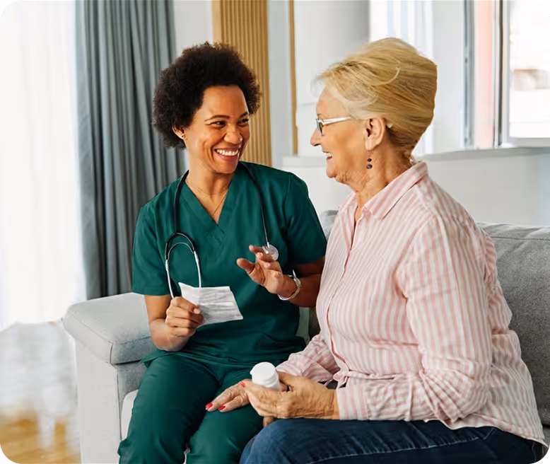 Smiling nurse in green scrubs discusses medication with a seated elderly woman at home.