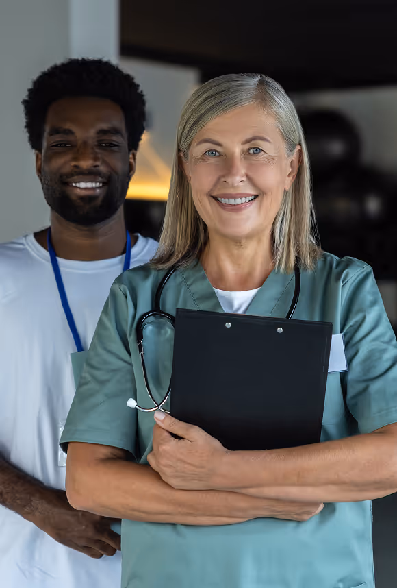 Smiling female healthcare worker in scrubs with clipboard and male colleague behind her in ID badge.