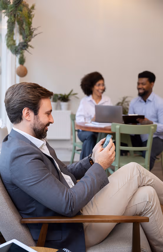 Man in suit sitting and using smartphone while two people chat at table in background.