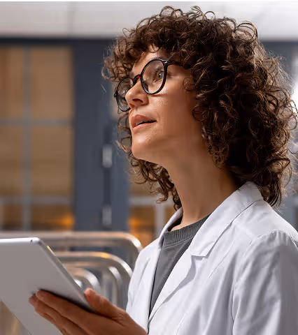 Curly-haired woman in glasses and white coat holding a tablet, looking thoughtfully upward.